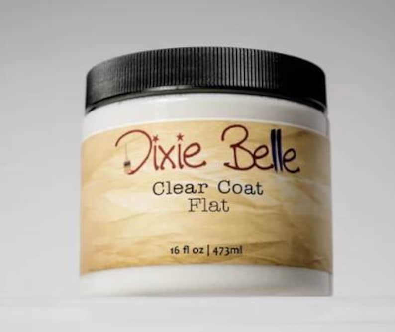Może przedstawiać: Biały plastikowy słoik z czarną zakrętką, posiadający beżową etykietę z napisem Dixie Belle Clear Coat Flat oraz 473ml. Pojemnik zawiera przezroczysty płyn wykończeniowy, a na etykiecie znajduje się mała grafika przedstawiająca pędzel.