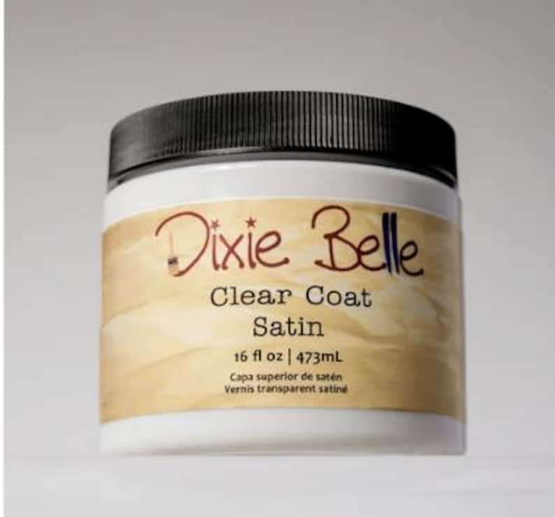 Może przedstawiać: Biały plastikowy słoik z czarną zakrętką zawierający Dixie Belle Clear Coat Satin. Etykieta zawiera nazwę marki, rodzaj produktu oraz objętość 473mL wydrukowaną czarnym tekstem na beżowym tle z ikoną pędzla.