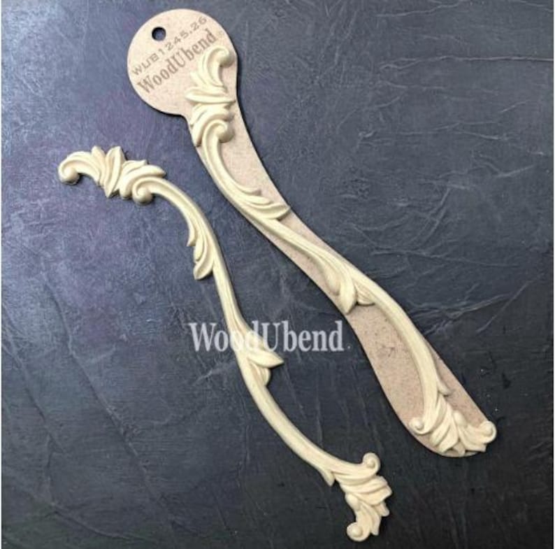 May include: Two cream-colored, decorative wood appliques with an ornate, scrolling leaf design. One is curved, the other is straight. The appliques are from WoodUbend, as indicated by the text on the tag and the product itself.