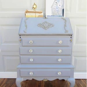 May include: A light blue desk with crystal knobs and gold accents. The desk has a drop-down writing surface and multiple drawers. The legs are curved with gold-colored feet. A small clock and artwork are on top of the desk.