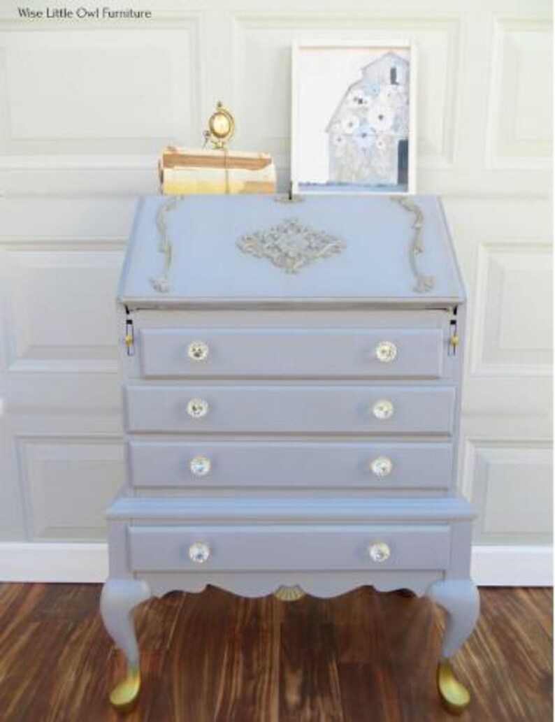 May include: A light blue desk with crystal knobs and gold accents. The desk has a drop-down writing surface and multiple drawers. The legs are curved with gold-colored feet. A small clock and artwork are on top of the desk.