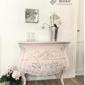 May include: A light pink painted chest of drawers with floral and scrollwork details. The chest has curved legs and a silver top. A silver metal leaf wall hanging and a metal bucket with flowers are on top.