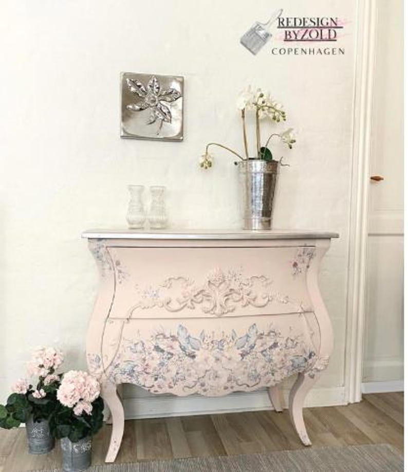 May include: A light pink painted chest of drawers with floral and scrollwork details. The chest has curved legs and a silver top. A silver metal leaf wall hanging and a metal bucket with flowers are on top.