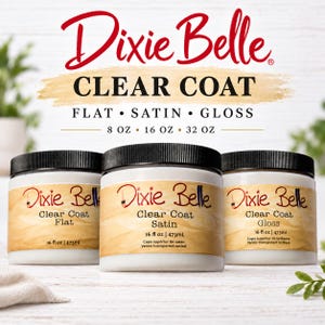 Może przedstawiać: Trzy białe plastikowe słoiki z czarnymi nakrętkami zawierające lakier Dixie Belle Clear Coat w wykończeniach Flat, Satin i Gloss. Każda etykieta zawiera nazwę marki, rodzaj wykończenia oraz objętość 16 fl oz lub 473 mL. Tekst: Dixie Belle.