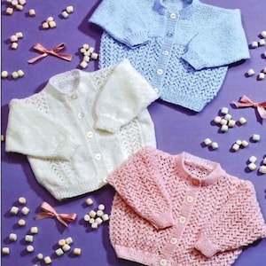 以下が含まれることがあります： パステルカラーのベビー用ニットカーディガン3着。水色、白、ピンクがあり、それぞれボタン留めと繊細な透かし模様が特徴です。紫色の表面に配置され、マシュマロとピンクのリボンが散りばめられています。
