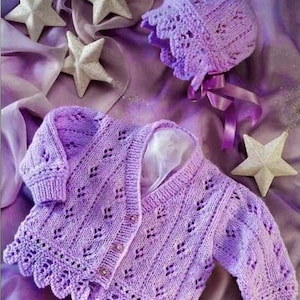 Op de afbeelding: Een lila gebreide babycardigan en muts. De cardigan heeft een geschulpte zoom en open details. De muts heeft een bijpassende kanten rand en strikjes. De set wordt getoond op een zachte, paarse stof met sterdecoraties.