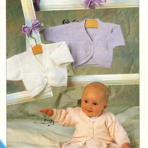 以下が含まれることがあります： ハンガーにかけられた、白とラベンダー色の2つのベビー用ニットカーディガン。赤ちゃんはピンクのカーディガンと白いドレスを着て、ベッドに座っています。