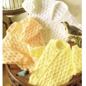 May include: Three knitted baby cardigans in white, yellow and peach with a textured stitch pattern. The cardigans are laid out on a wooden table with a bird figurine.