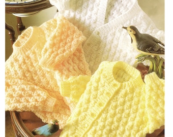 May include: Three knitted baby cardigans in white, yellow, and peach with a textured stitch pattern. The cardigans are laid out on a wooden table with a bird figurine.