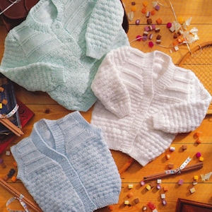 May include: Three hand-knitted baby sweaters in soft pastel colors: a light blue cardigan, a white cardigan, and a light blue vest. The sweaters are made of textured yarn and are displayed on a wooden surface with colorful candies.
