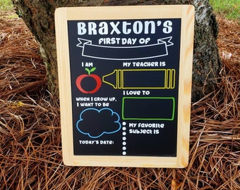 Puede incluir: Una pizarra negra con marco de madera, decorada con una manzana roja, un crayón amarillo y una nube azul. La pizarra tiene el texto "Braxton's First Day Of" en la parte superior e incluye espacios para escribir la edad del niño, el nombre del maestro, la materia favorita y la fecha de hoy.