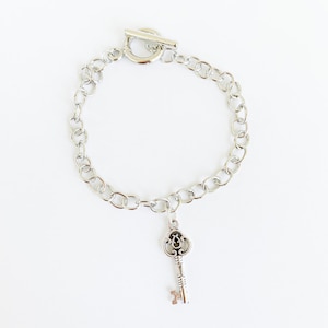 May include: Silver-toned bracelet with a key charm. The bracelet features a chain link design and a toggle clasp. The key charm is detailed with intricate patterns. The jewelry is displayed on a white background.