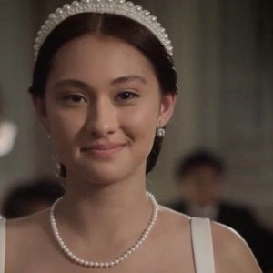 Puede incluir: Una mujer con un collar de perlas, pendientes y una tiara. Sonr&iacute;e, y el fondo est&aacute; borroso, lo que sugiere un evento formal. La iluminaci&oacute;n es suave y el tono general es elegante y refinado.