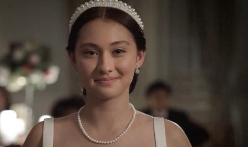 Puede incluir: Una mujer con un collar de perlas, pendientes y una tiara. Sonr&iacute;e, y el fondo est&aacute; borroso, lo que sugiere un evento formal. La iluminaci&oacute;n es suave y el tono general es elegante y refinado.