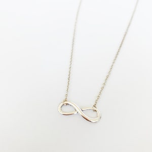 May include: A silver-colored necklace featuring an infinity symbol pendant. The pendant is connected to a delicate chain. The necklace is displayed against a white background, highlighting its simple elegance.
