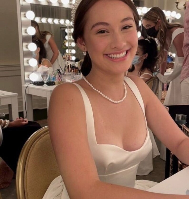 Puede incluir: Una mujer con un vestido de sat&eacute;n blanco con escote cuadrado y tirantes finos. Tambi&eacute;n lleva un collar de perlas y pendientes. El fondo muestra un espejo con luces y otras personas con vestidos blancos.