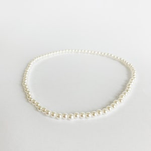 Puede incluir: Un delicado collar de perlas de color blanco roto. El collar est&aacute; compuesto por numerosas perlas peque&ntilde;as y redondas ensartadas, formando un c&iacute;rculo completo. Las perlas tienen un brillo sutil y el collar descansa sobre un fondo blanco.