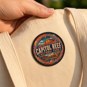 May include: A circular embroidered patch featuring a landscape illustration of Capitol Reef National Park with red rock formations and a road, with the text CAPITOL REEF NATIONAL PARK in blue, attached to a cream-colored canvas tote bag.