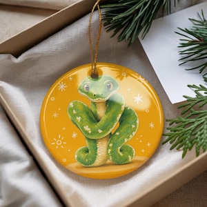 May include: A round ceramic ornament featuring a green snake illustration on a vibrant yellow background with white star accents. The ornament has a glossy finish and is suspended by a thin gold string, showcasing a detailed cartoon-style snake.