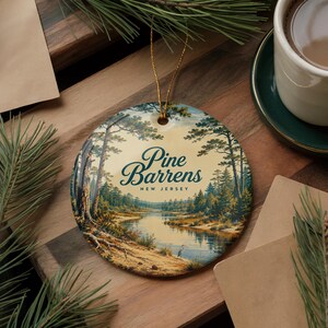 May include: A round ceramic ornament featuring a printed landscape of the Pine Barrens in New Jersey, depicting a river flowing through a forest with tall pine trees and a small bird, suspended by a thin gold string for hanging display.