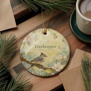 May include: A round ceramic ornament with a bird perched on a branch, surrounded by flowers and butterflies. The ornament has the word "Treehopper" printed on it. A gold string is attached for hanging. The background is a soft, pastel color palette.