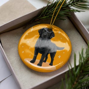 May include: A round, yellow Christmas ornament featuring a black Labrador Retriever with a gray overlay. The ornament has a gold string for hanging and is decorated with white stars and sunbursts. It is presented in a gift box.