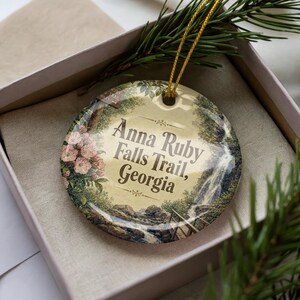 May include: A round ceramic ornament featuring a scenic illustration of Anna Ruby Falls Trail, Georgia. The design includes a waterfall, lush greenery, and pink flowers. The ornament has a gold string for hanging and is presented in a gift box.