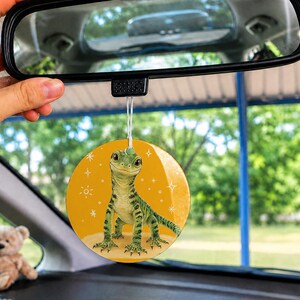May include: A round car air freshener hanging from a rearview mirror, featuring a green lizard illustration on a yellow background with white star accents. The lizard has large eyes and a textured body, suspended by a thin white string.