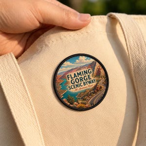 May include: A circular embroidered patch featuring a landscape illustration of Flaming Gorge with a winding road, blue water, and rocky cliffs. The text reads FLAMING GORGE SCENIC BYWAY All-American Road. The patch is attached to a beige canvas tote bag.