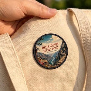May include: A circular embroidered patch featuring a landscape illustration of Hells Canyon Scenic Byway with a winding road, river, and mountains. The text reads Hells Canyon Scenic Byway All-American Road. The patch is attached to a beige canvas tote bag.