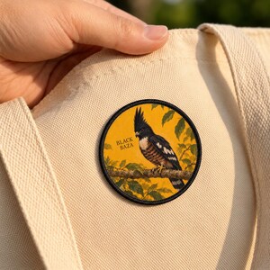 May include: A circular embroidered patch featuring a black and brown Black Baza bird perched on a branch against a yellow background with green leaves. The text BLACK BAZA is printed in black on the left side of the patch.