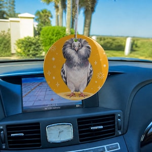 May include: A circular car air freshener hanging from a rearview mirror, featuring a graphic illustration of a grey Jacobin pigeon with a large, fluffy feathered hood against a solid mustard yellow background with small white star and sun icons.