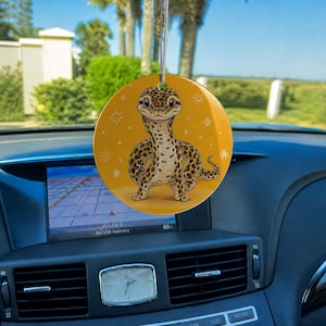 May include: A circular hanging car ornament featuring a yellow background with a central illustration of a brown and tan spotted leopard gecko. The gecko is depicted in a standing position with a smiling expression, surrounded by small white star icons.