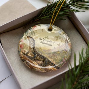 May include: A round ceramic ornament featuring a Ridge-tailed Monitor lizard illustration. The ornament has a gold string for hanging and is presented in a gift box with a sprig of greenery.