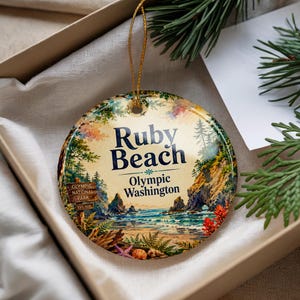 May include: A round ceramic ornament featuring a watercolor illustration of Ruby Beach in Olympic National Park, Washington. The text reads Ruby Beach, Olympic Washington, and Olympic National Park Est 1938. It hangs from a gold string.