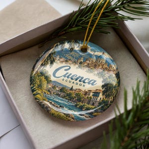 May include: A round ceramic ornament featuring a colorful illustration of the city of Cuenca, Ecuador, with a river, historic buildings, and a cathedral dome. The text Cuenca Ecuador is printed in the center. It includes a gold hanging string.