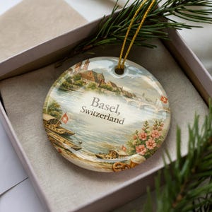 May include: A round ceramic ornament featuring a scenic view of Basel, Switzerland, with the city's name printed on it. The ornament has a gold string for hanging and is presented in a gift box. The image depicts boats, buildings, and a river.