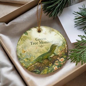 May include: A round ceramic ornament featuring a green tree monitor lizard on a tree branch. The ornament has the text "Green Tree Monitor" and is decorated with butterflies and flowers. It is presented in a gift box.
