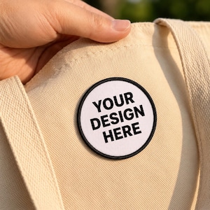 May include: A circular patch with a white background and a black border attached to a beige canvas tote bag. The patch features the black text YOUR DESIGN HERE in a bold, sans-serif font, centered within the circular frame on the fabric.