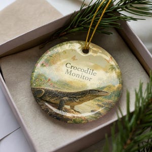 May include: A round ceramic ornament featuring a Crocodile Monitor illustration. The ornament has a gold string for hanging and is presented in a gift box. The background shows a landscape with butterflies and the text "Crocodile Monitor".