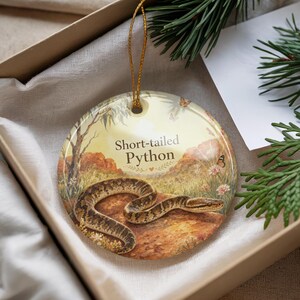 May include: A round ceramic ornament featuring a detailed illustration of a short-tailed python in a natural landscape. The ornament has a gold string for hanging and the text "Short-tailed Python".