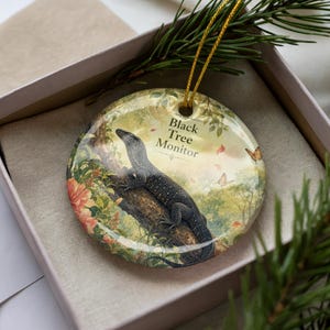 May include: A round ceramic ornament featuring a Black Tree Monitor lizard perched on a tree branch. The ornament has a gold string for hanging and is set in a gift box. The background includes flowers, butterflies, and the text "Black Tree Monitor."