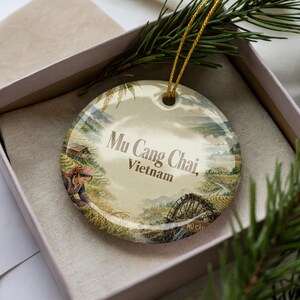 May include: A round ceramic ornament featuring a scenic illustration of Mu Cang Chai, Vietnam. The ornament has a gold string for hanging and is presented in a gift box. The illustration depicts a woman in traditional clothing and a water wheel.