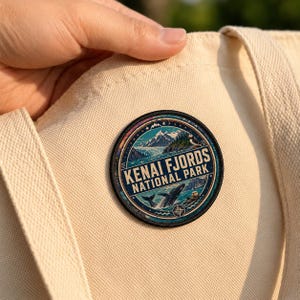 May include: A circular embroidered patch featuring a landscape of Kenai Fjords National Park, including mountains, a glacier, a whale, and a puffin, with the text "KENAI FJORDS NATIONAL PARK" in white, attached to a cream-colored canvas tote bag.