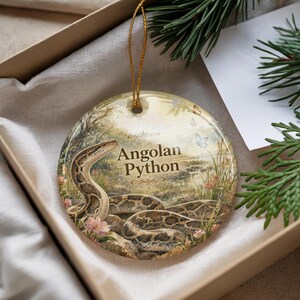 May include: A round ceramic ornament featuring an Angolan Python in a naturalistic setting. The ornament has a gold string for hanging and is presented in a gift box. The text "Angolan Python" is printed on the ornament.