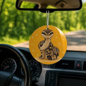 May include: A circular car air freshener hanging from a rearview mirror, featuring a mustard yellow background with a cartoon illustration of a brown and tan ball python snake surrounded by small white star and sunburst graphic accents.