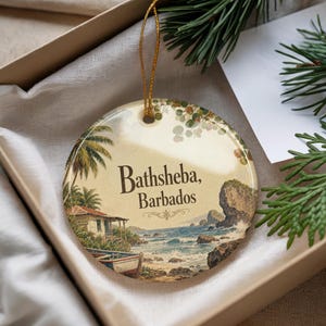 May include: Round ceramic ornament featuring a coastal scene with the text "Bathsheba, Barbados." The design includes a beach, ocean, palm trees, and a small boat. The ornament is in a gift box with a gold string for hanging.