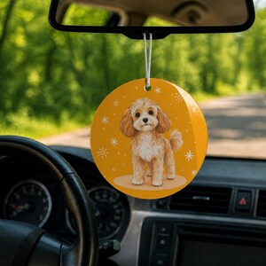 May include: A circular car ornament hanging from a rearview mirror, featuring a mustard yellow background with a detailed illustration of a light brown curly-haired puppy and small white snowflake patterns printed on the surface.