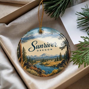 May include: A round ceramic ornament featuring a landscape illustration of Sunriver, Oregon, with a blue river, pine trees, and snow-capped mountains. The text Sunriver OREGON is printed in dark blue script and block letters. It has a gold string.