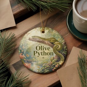 May include: Round ceramic ornament featuring an olive python coiled on a tree branch. The background depicts a serene landscape with a river, flowers, and butterflies. The text "Olive Python" is displayed in a decorative font.